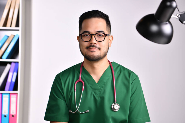 Портрет молодого врача-азиата в хирургической форме, слушающего и обсуждающего с пациентом онлайн, делающего видеозвонок, смотрящего в камеру. - Фото, изображение