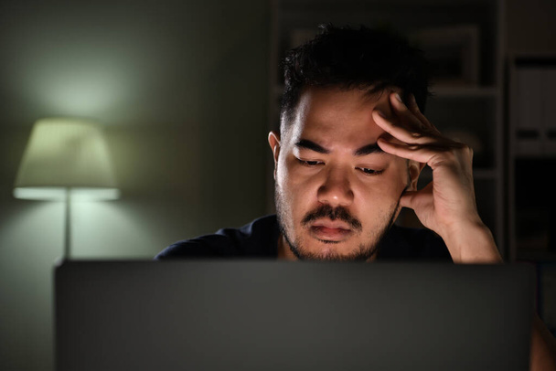 Feeling stressful and depressed. Asian man feeling looking unhappy, stressed and tired while using computer working late at night at home - Фото, изображение