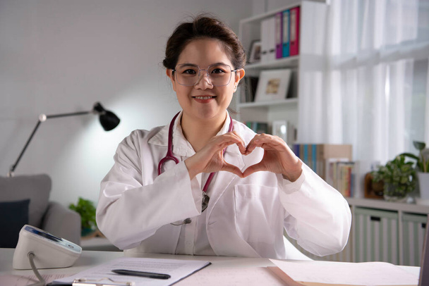 Портрет молодої вродливої азіатки-жінки-лікаря робить серцевий знак, дивлячись на камеру в медичному кабінеті, маючи онлайн-консультації з пацієнтом. Молода жінка - лікар з стетоскопом робить відеодзвінок. - Фото, зображення