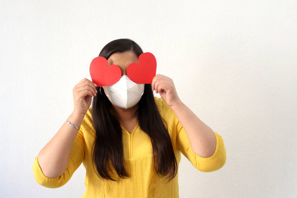 Latin adult woman with a yellow blouse and a big red heart shows her enthusiasm for Valentine's Day of Love and Friendship with a face mask in the new normality due to the Covid-19 pandemic - Foto, Imagen