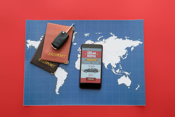Teléfono móvil con aplicación de alquiler de coche abierto, pasaportes, llave y mapa sobre fondo rojo - Foto, imagen