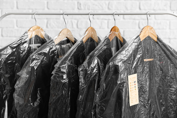 Bastidor con chaquetas negras limpias en bolsas de plástico sobre fondo de ladrillo blanco, primer plano - Foto, Imagen