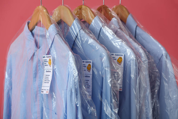 Rack with clean blue shirts in plastic bags on pink background, closeup - Foto, Imagem