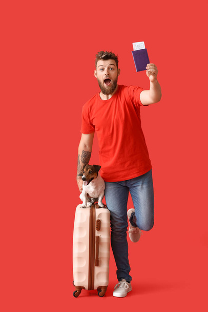 Überraschter junger Mann mit Pass, niedlichem Hund und Koffer auf rotem Hintergrund - Foto, Bild