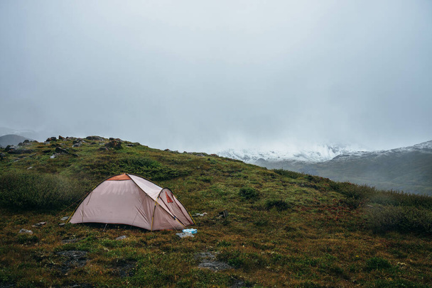 Scenic alpine landscape with tent on green hill among rocks. Beautiful snow-capped mountains in low clouds. Atmospheric scenery with tent in highlands in overcast weather. High mountains covered snow. - Foto, Imagen