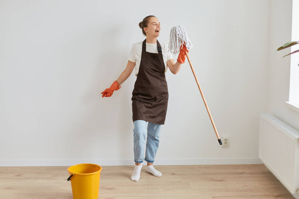 Full length portrait of happy woman wearing orange rubber gloves, brown apron and jeans washing floor at home, singing in mop, having fun while cleaning her flat. - 写真・画像