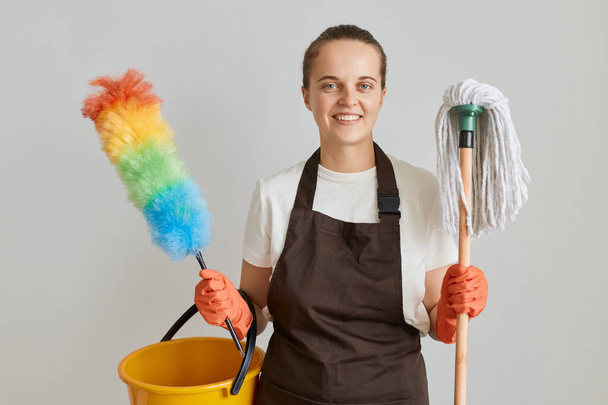 Indoor shot of smiling woman housekeeper wearing brown apron and white t shirt cleaning her apartment, holding mop, ppduster and bucket and being going to wash her house. - Фото, зображення