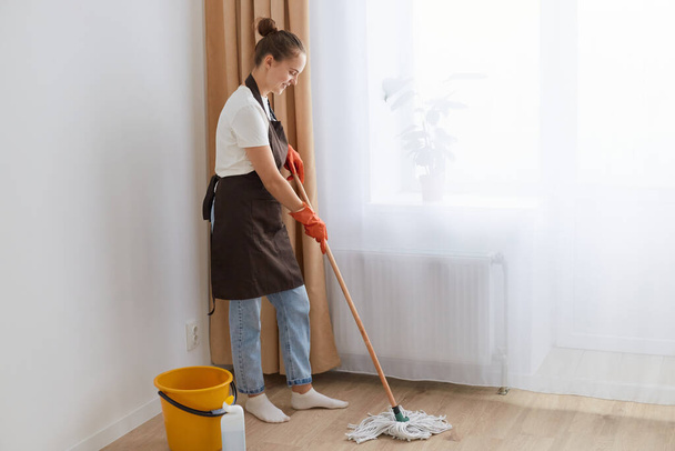 Full length portrait of woman wearing orange rubber gloves, brown apron and jeans washing floor at home, holding mop, looking down, doing her household chores. - Fotografie, Obrázek