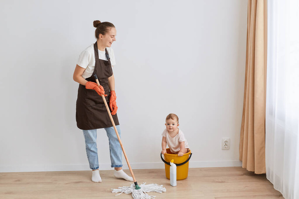 Full length portrait of laughing woman wearing orange rubber gloves, brown apron and jeans washing floor at home, looking at her funny infant daughter in yellow bucket. - Photo, Image