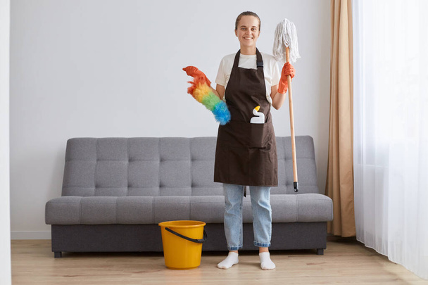 Zdjęcie uśmiechniętej kobiety w brązowym fartuchu, dżinsach i białej koszuli sprzątającej mieszkanie, trzymającej detergent w kieszeni, patrzącej w kamerę ze szczęśliwym wyrazem twarzy, cieszącej się sprzątaniem. - Zdjęcie, obraz