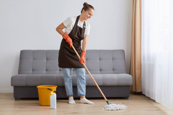 Full length portrait of woman in good mood wearing brown apron, jeans and white t shirt cleaning her house, washing floor near sofa, likes doing household chores. - Photo, Image