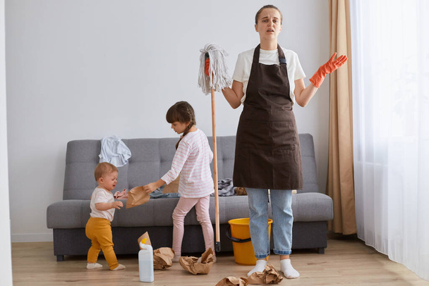 Indoor shot of stresses depressed woman, young mother wearing brown apron, jeans and white t shirt cleaning her house, children making mess, female looks nervous. - Fotó, kép