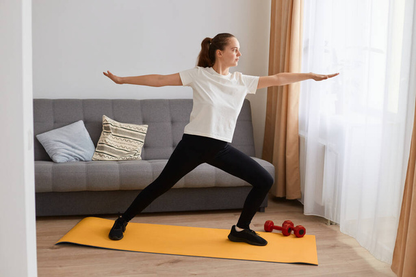 Indoor shot of attractive Caucasian young woman practicing yoga on exercise mat in living room while resting at home, working out, female wearing sportswear. - Foto, immagini