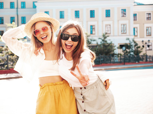 Two young beautiful smiling hipster female in trendy summer clothes.Sexy carefree women posing on the street background in hat. Positive pure models having fun at sunset, hugging - Foto, Bild