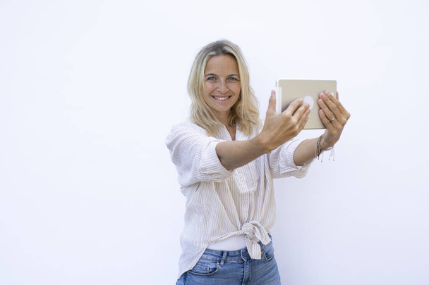 beautiful young blond woman wearing blue jeans having video call using silver tablet in front of white background - Foto, afbeelding