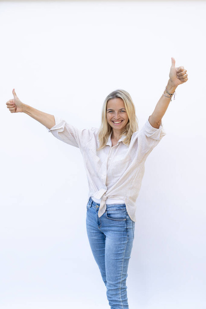 blond woman with blue jeans and blue eyes is posing in front of white background and showing thumbs up  - 写真・画像
