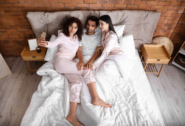 Homme avec deux belles femmes prenant selfie au lit. Concept polyamory - Photo, image