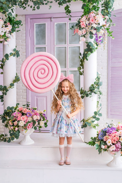 girl in a doll dress and with a bow on her head holds a large candy lollipop on a stick - Photo, Image