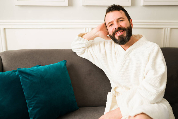 Portrait of a cheerful latin man making eye contact and smiling while relaxing on a white robe at home after a shower - Photo, Image