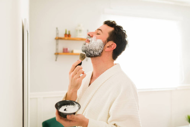 Vista laterale di un uomo barbuto che si guarda allo specchio e mette la crema da barba per rimuovere i peli sul viso - Foto, immagini