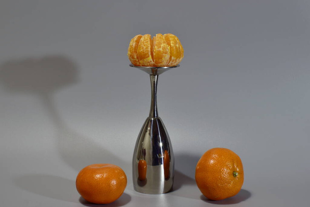 three tangerines, a metal glass and shadow from them on a gray background - Фото, изображение
