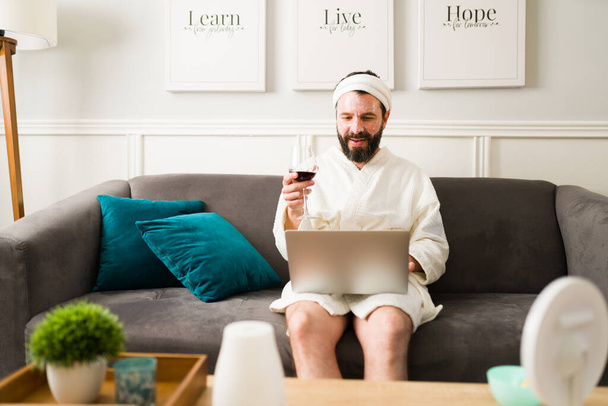 Relaxed man drinking wine and smiling while doing online shopping on the laptop. Hispanic man having a spa day at home  - Photo, Image