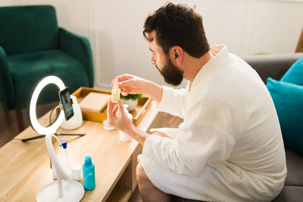 Vista posteriore di un uomo che utilizza un siero e prodotti per la cura della pelle durante le riprese di un video con il suo smartphone e una luce anulare - Foto, immagini