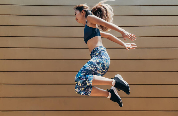 Atleta femenina seria haciendo ejercicio al aire libre. Mujer asiática en ropa deportiva haciendo saltos de altura como parte de su rutina de ejercicios. - Foto, Imagen