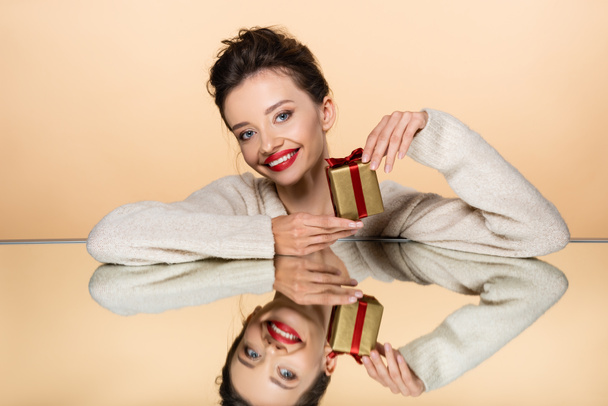 Smiling woman with red lips holding present near mirror isolated on beige  - Fotografie, Obrázek