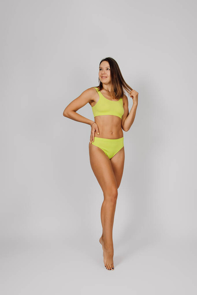 Brunette shows off slender figure. Young beautiful Caucasian woman in yellow swimsuit or bikini poses on light isolated background, stands in elegant pose and holds her hair with her hands - Photo, Image