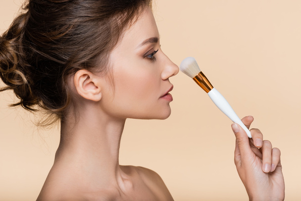 Side view of brunette woman holding cosmetic brush near nose isolated on beige  - Zdjęcie, obraz