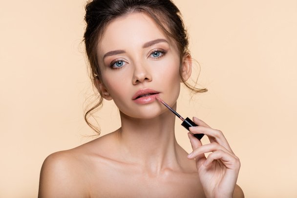 Young woman with naked shoulders applying lip gloss isolated on beige  - Foto, afbeelding