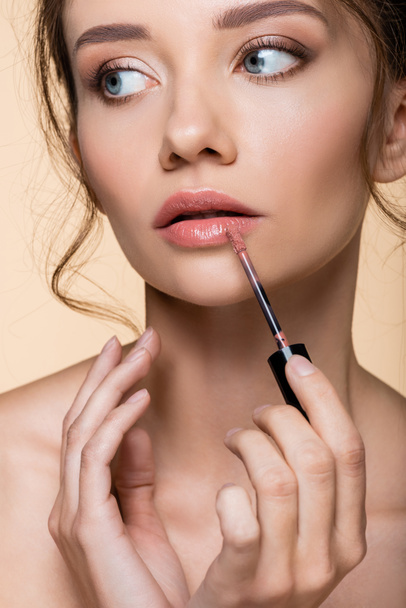 Portrait of pretty woman applying lip gloss and looking away isolated on beige  - Zdjęcie, obraz