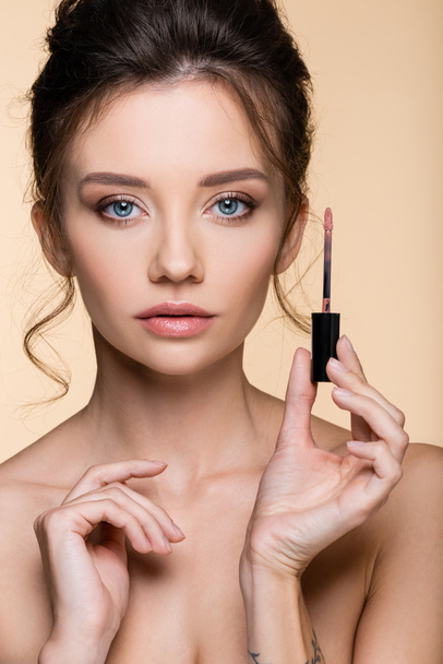 Retrato de modelo bonito sosteniendo cepillo de brillo de labios y mirando a la cámara aislada en beige  - Foto, Imagen