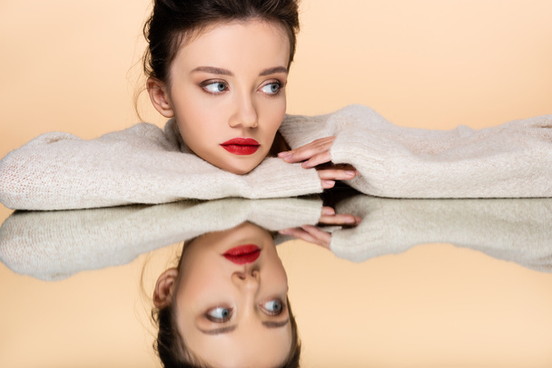 Pretty model with red lips wearing sweater and looking away near mirror isolated on beige  - Foto, afbeelding