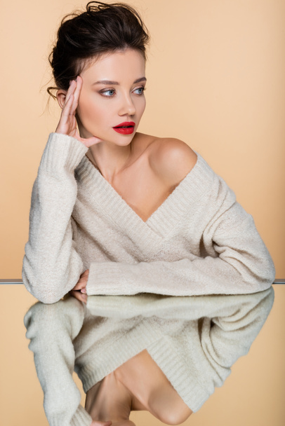 Trendy model in sweater with naked shoulder looking away near mirror isolated on beige  - Foto, afbeelding