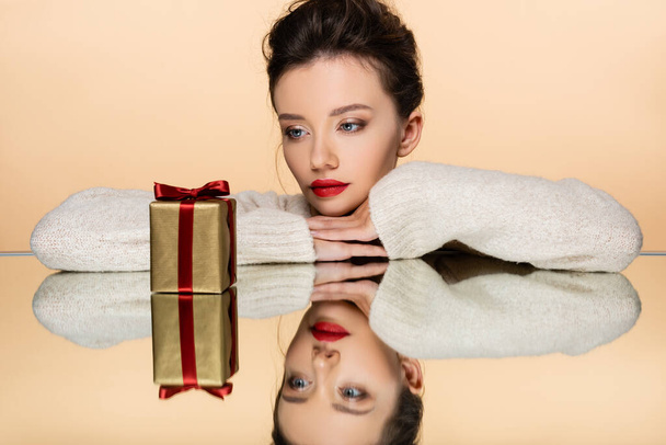 Pretty woman in sweater looking at gift on mirror isolated on beige  - 写真・画像