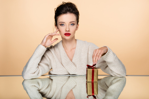 Stylish woman in warm sweater looking at camera near gift on mirror isolated on beige  - Foto, immagini