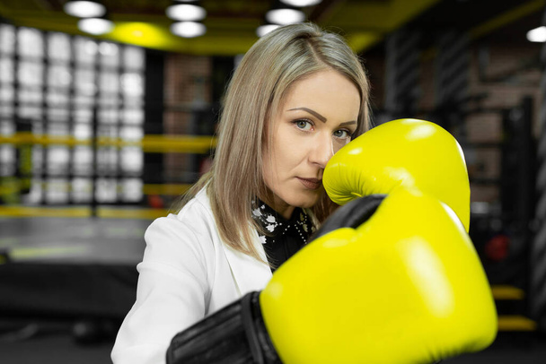 Determined, stylish businesswoman in yellow boxing gloves throws a punch at the camera against the backdrop of a boxing ring - Fotó, kép