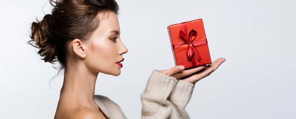 Vista lateral de modelo morena en suéter mirando caja de regalo con cinta aislada en blanco, banner  - Foto, Imagen