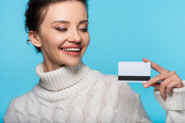 Mujer sonriente en jersey de punto mirando la tarjeta de crédito aislada en azul - Foto, imagen