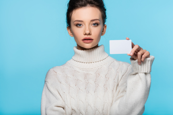 Mladá žena v pleteném svetru drží prázdnou kartu a dívá se na kameru izolované na modré - Fotografie, Obrázek