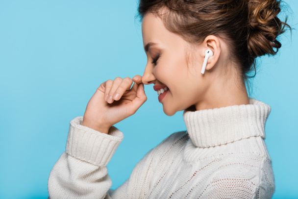 Πλευρική άποψη της χαρούμενης γυναίκας σε πλεκτό πουλόβερ και ακουστικό απομονώνονται σε μπλε - Φωτογραφία, εικόνα