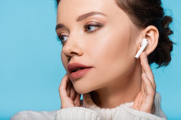 Young model in sweater using wireless earphone isolated on blue - Фото, изображение