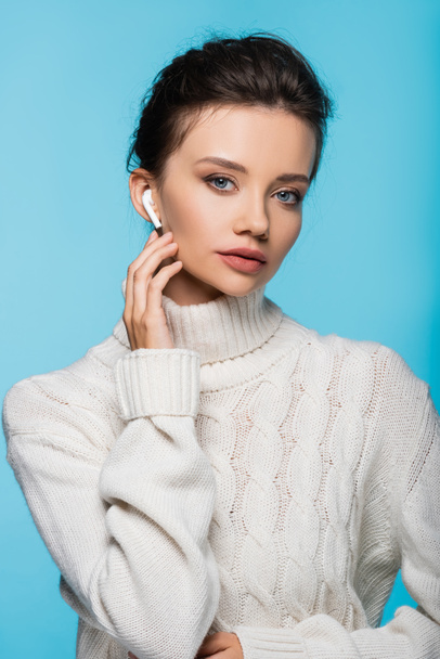 Model in white sweater using earphone and looking at camera isolated on blue - Фото, изображение