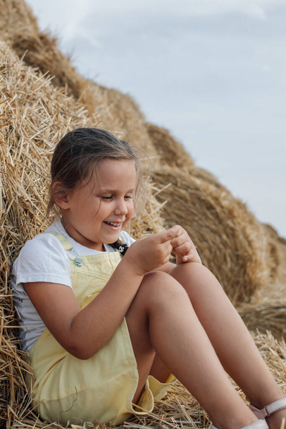 Portrait of cute girl holding piece of hay in hands smiling with teeth and looking at hands sitting on haystack with another stack behind back with dozens of haystacks in background. Away from city - Photo, Image