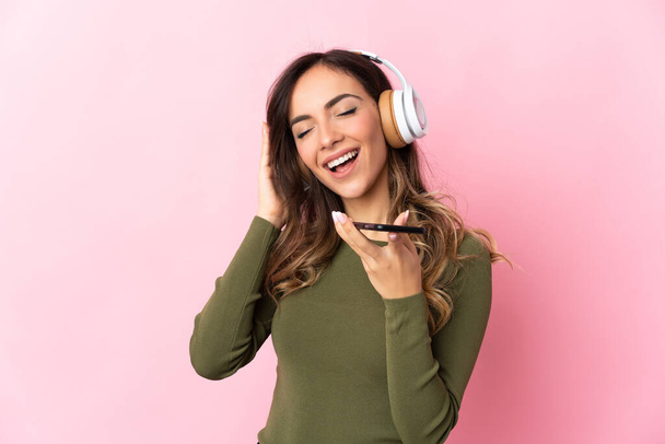 Junge kaukasische Frau isoliert auf rosa Hintergrund Musik hören mit einem Handy und singen - Foto, Bild
