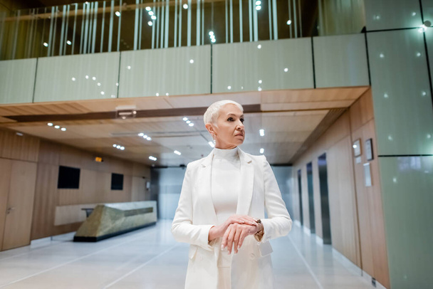 mature businesswoman in white trendy clothes looking away in lobby of modern hotel - Foto, afbeelding