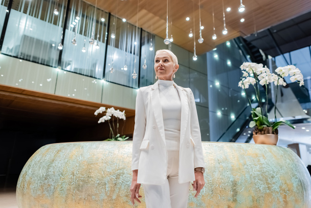 blonde senior businesswoman in white clothes standing near orchids in luxury hotel - Foto, Imagen
