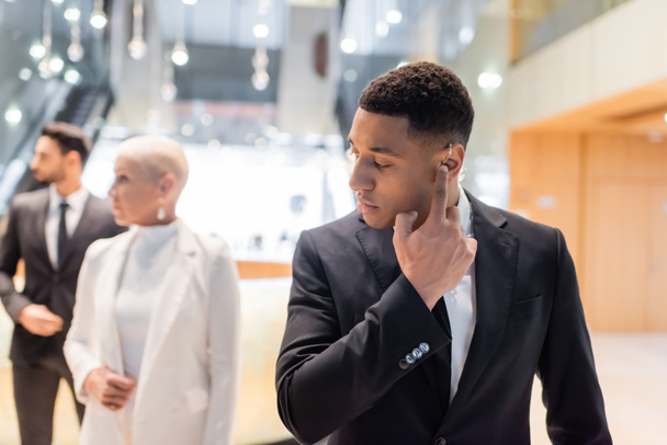 african american bodyguard adjusting earpiece near mature businesswoman with bi-racial guard on blurred background - Photo, Image